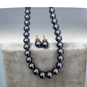 May include: A dark gray pearl necklace and matching stud earrings set. The necklace and earrings feature large, round, lustrous pearls. The earrings have gold-colored posts. The jewelry is displayed on a light gray, textured surface.