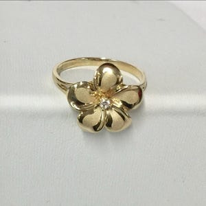 May include: A gold-colored ring featuring a flower design. The flower has five petals and a small, clear gemstone in the center. The ring is set on a simple band. The ring is likely made of gold or gold-plated metal.