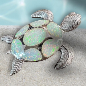 May include: A silver-toned turtle figurine with a clear shell filled with oval-shaped opalescent stones. The stones have a mix of green, white, and orange hues. The turtle's head, flippers, and shell edges are detailed with textured patterns.