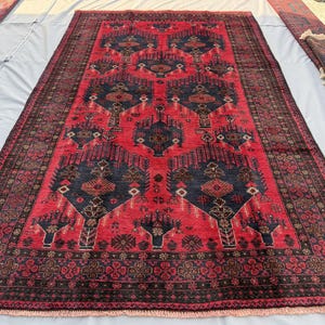 May include: A large, rectangular red rug with a detailed geometric pattern in navy blue and brown. The rug has a dark border and intricate central designs. Additional rolled rugs are visible on the sides.