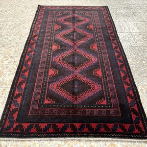 Pode incluir: Um tapete retangular com um design geométrico central em vermelho e castanho escuro. O tapete tem uma borda preta com detalhes vermelhos e azuis. O tapete é feito de material tecido, adequado para uma sala de estar ou quarto.