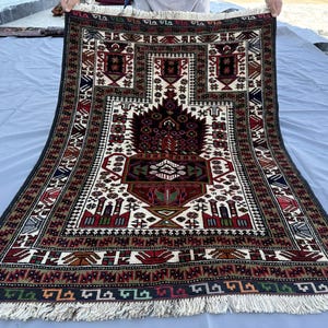 May include: A rectangular, hand-woven rug with a central design featuring a tree-like motif in shades of red, brown, and black. The rug has a cream-colored background with geometric patterns and a fringed edge. The border is dark blue with repeating patterns.