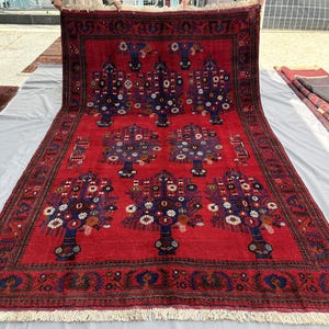 May include: A large, rectangular red rug with a detailed floral pattern. The design features stylized trees with blue vases and colorful blossoms. The rug has a dark border and a fringed edge, showcasing traditional craftsmanship.