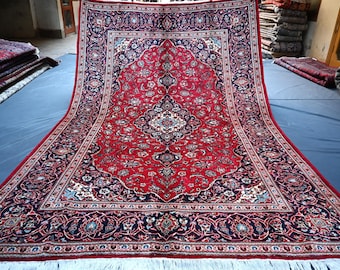 Puede incluir: Una alfombra persa roja, azul y blanca con intrincados patrones florales. La alfombra tiene un borde con flecos y está doblada por la mitad.