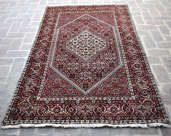 Op de afbeelding: Een traditioneel Perzisch tapijt met een rood, wit en zwart kleurenschema. Het tapijt heeft een centraal medaillon met ingewikkelde bloem- en geometrische patronen. Het tapijt heeft een gefranst randje.