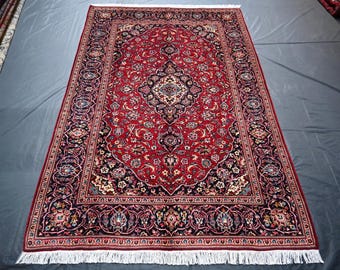 Puede incluir: Una alfombra persa roja, azul y crema con un patrón floral. La alfombra tiene un borde con flecos.