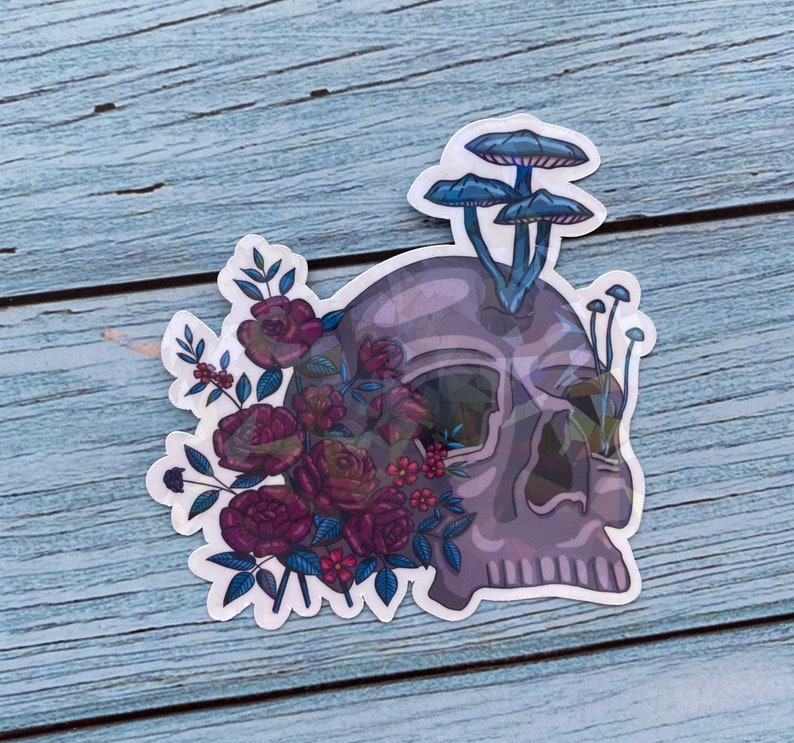 May include: A sticker featuring a purple skull with a bouquet of dark pink roses and blue mushrooms growing from the top of the skull.