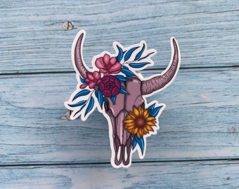 May include: A sticker featuring a floral skull with pink, purple, and yellow flowers. The skull has horns and is surrounded by blue leaves.