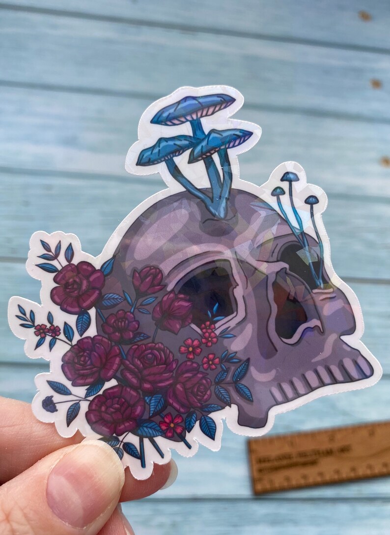 May include: A sticker featuring a purple and blue skull with a cluster of blue mushrooms growing from the top. The skull is surrounded by a bouquet of dark red roses and small pink flowers.