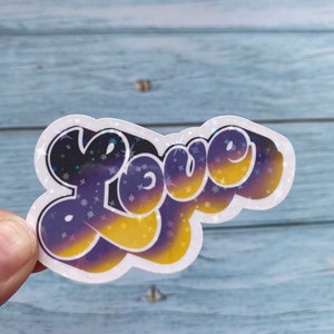 May include: A sticker featuring the word "Love" in a retro, bubbly font. The letters are a gradient of purple, black, and yellow with a glittery finish.