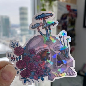 May include: A holographic sticker featuring a skull with a cluster of purple roses and three blue mushrooms growing from the top of the skull.