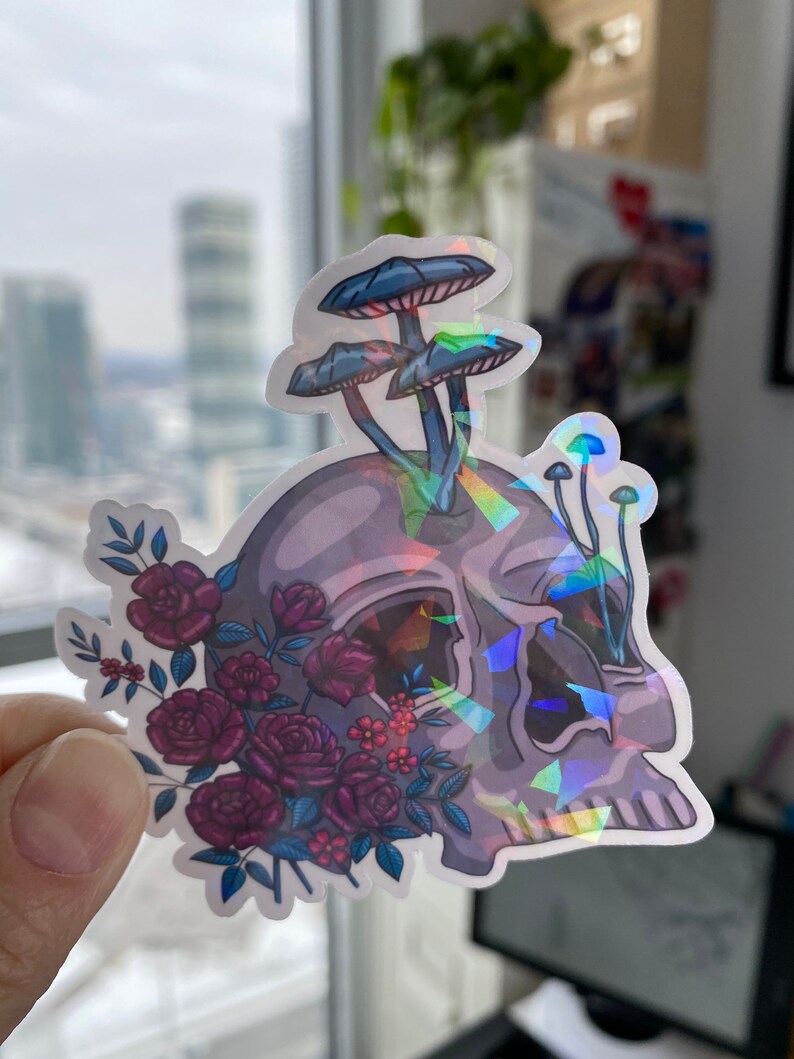 May include: A holographic sticker featuring a skull with a cluster of purple roses and three blue mushrooms growing from the top of the skull.
