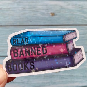 May include: A sticker featuring a stack of three books. The top book is blue with the word "READ" on it. The middle book is pink with the word "BANNED" on it. The bottom book is purple with the word "BOOKS" on it. The books are surrounded by a white border and a glittery background.
