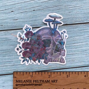 May include: A sticker featuring a skull with a floral design and mushrooms growing from the top. The skull is a light gray color with a purple and blue gradient. The flowers are a deep purple and red color with green leaves. The mushrooms are a light blue color with white tops.