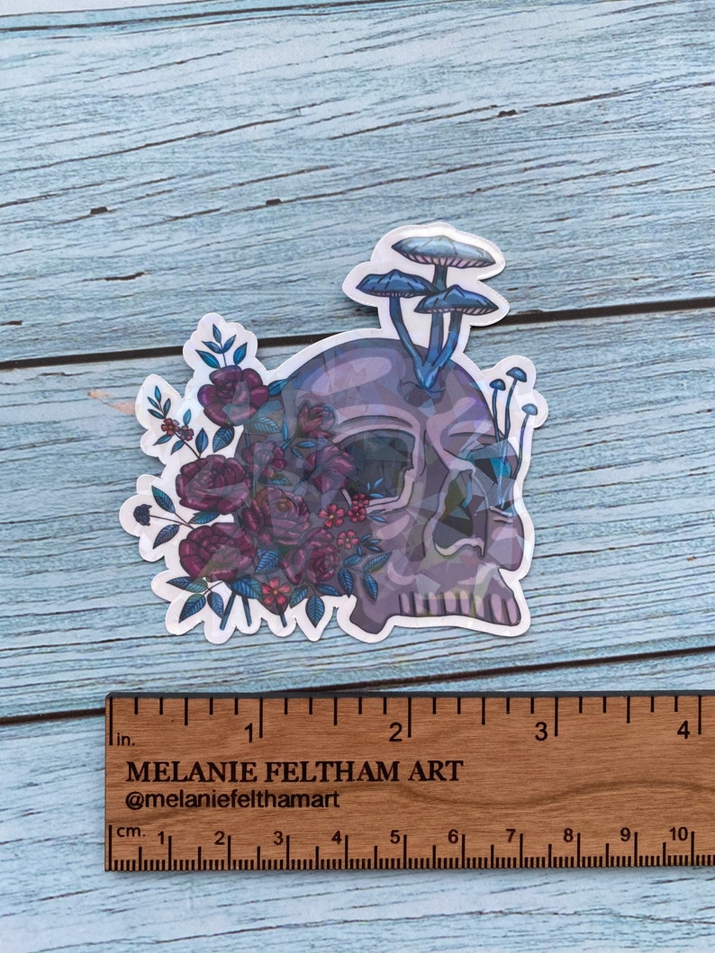 May include: A sticker featuring a skull with a floral design and mushrooms growing from the top. The skull is a light gray color with a purple and blue gradient. The flowers are a deep purple and red color with green leaves. The mushrooms are a light blue color with white tops.