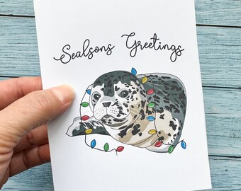 May include: A Christmas card featuring a cartoon seal wearing a string of colorful Christmas lights. The card reads "Seasons Greetings".