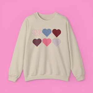 May include: A beige sweatshirt featuring six heart-shaped designs in various patterns and colors. The hearts include red, blue, and pink, with patterns like dots, stripes, and plaid. The sweatshirt has long sleeves and a crew neck.