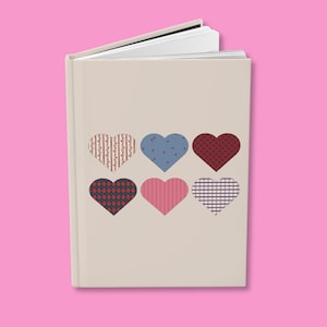 May include: A beige journal with six patterned hearts in various colors. The hearts include designs like stripes, polka dots, and plaid. The journal is open, revealing white pages, and set against a pink background.