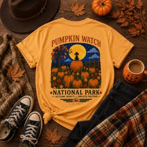 May include: A mustard yellow t-shirt featuring a graphic print with the text PUMPKIN WATCH NATIONAL PARK AUTUMN NIGHTS ENDLESS WAITING EST 1986. The illustration depicts a dog sitting on a large pumpkin under a full moon in a pumpkin patch.
