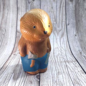Puede incluir: Una figura marrón y beige de un oso con pantalones cortos azules y una hacha. El oso tiene pequeños ojos negros y está de pie. La figura está hecha de un material duro.