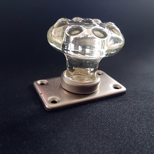Peut inclure: Bouton de porte en verre transparent avec un dessus décoratif et arrondi. Le bouton est monté sur une base rectangulaire en métal argenté avec quatre trous de vis. Le fond est de couleur sombre et unie.