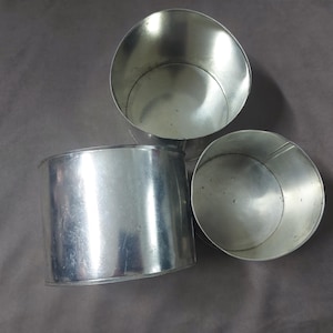 May include: Three open, silver-colored metal containers. One is cylindrical, and the other two are round with a slightly tapered shape. The containers are empty and have a reflective surface, suggesting they are made of tin or a similar metal.