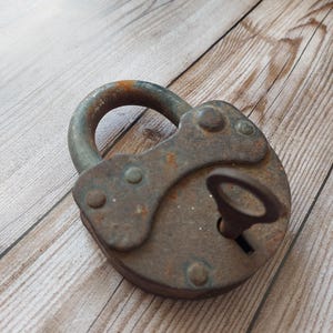May include: An old, rusty padlock with a key in the keyhole. The metal lock has a rounded design and a curved handle. The padlock shows signs of age and wear, resting on a wooden surface.