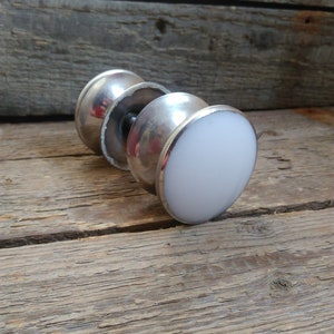 May include: A silver-toned door knob with a white, rounded end. The knob has a cylindrical shape with a central screw and is set against a weathered wooden surface. The design is simple and classic.
