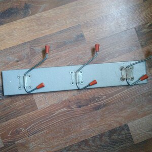 Puede incluir: Un perchero de metal plateado con cuatro ganchos. Los ganchos tienen puntas de plástico naranja. El perchero está montado en una pared de madera.