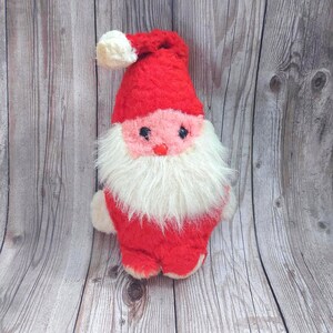 May include: A plush Santa Claus doll with a red hat and suit, white beard, and pink face. The doll has a white pom-pom on the hat and small white hands and feet. The doll is a Christmas decoration.