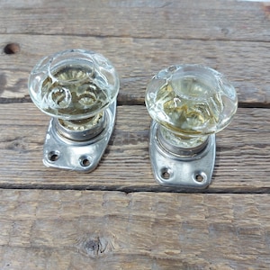 Puede incluir: Dos pomos de puerta antiguos de cristal transparente, con diseño facetado y tinte amarillento. Cada pomo está montado sobre una placa trasera de metal plateado con dos orificios para tornillos. Sobre una superficie de madera envejecida.