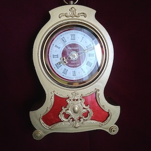 May include: A gold-colored clock with a red face and ornate details. The clock face has Roman numerals and the text "Zenit" and "Kварц" in Cyrillic script. The clock is on a dark red background.