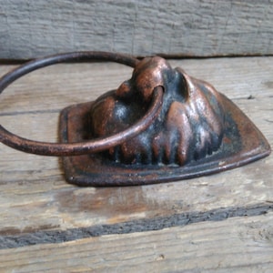 Könnte beinhalten: Ein rustikaler, antiker Türklopfer mit einem Ringgriff. Der Türklopfer ist aus Metall und hat eine dunkelbraune Oberfläche mit kupferfarbenen Akzenten. Der Türklopfer ist in Form eines Gesichts mit einem erhabenen, strukturierten Design gestaltet.