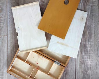 May include: A wooden artist's paint box with a hinged lid, open to reveal compartments. A wooden palette with a thumb hole and two wooden panels are next to the box. The wood has a natural finish.