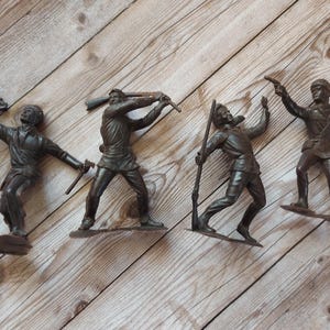 May include: Four brown plastic toy soldiers in various action poses, each holding a weapon. One figure holds a knife, another a rifle, and two others hold pistols. The figures are on a wooden surface.