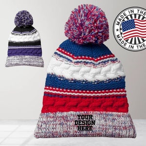 May include: A red, white, and blue striped knit beanie with a pom pom on top. The beanie has a white band with the text "YOUR DESIGN HERE" in black. The beanie is made in the USA.