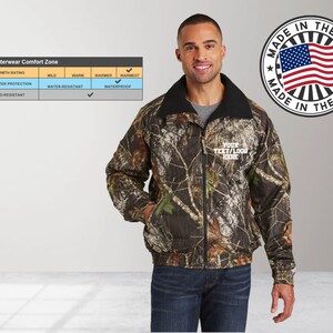 May include: A camouflage print jacket with a black fleece lining. The jacket has a zipper closure and two pockets. The jacket is water-resistant and wind-resistant. The jacket is made in the USA. The text "YOUR TEXT/LOGO HERE" is embroidered on the jacket.