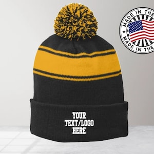 May include: Black and gold striped beanie with a black pom pom on top. The beanie has a fold-over cuff with the text "YOUR TEXT/LOGO HERE" embroidered in white.