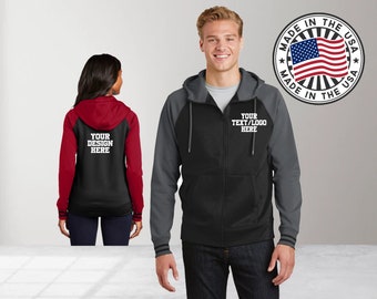 May include: Two black and grey color block zip-up hoodies with contrasting red and grey sleeves. The hoodie on the left has the text "YOUR DESIGN HERE" on the back. The hoodie on the right has the text "YOUR TEXT/LOGO HERE" on the front. Both hoodies have a white zipper. The image also includes a circular graphic with the text "MADE IN THE USA" and a red, white, and blue American flag.