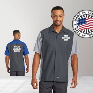 May include: Two men wearing gray and blue work shirts with short sleeves. The shirts have a contrasting color panel on the back and a white embroidered logo on the front. The logo says "YOUR TEXT/LOGO HERE". The shirt on the left has a blue panel and the shirt on the right has a gray panel. The shirts are made in the USA.