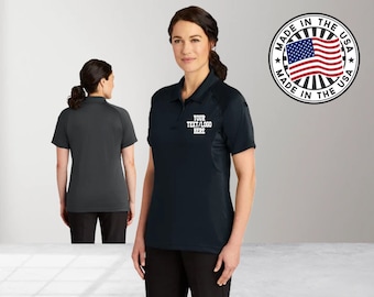 May include: Black polo shirt with a white embroidered logo that says "YOUR TEXT/LOGO HERE". The shirt has a mesh panel on the side. The model is wearing black pants. The image also includes a circular logo with the text "MADE IN THE USA" and a red, white, and blue American flag.