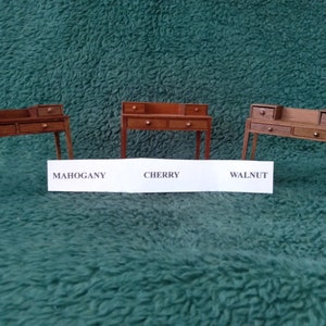 May include: Three miniature wooden desks in mahogany, cherry, and walnut finishes. Each desk has four drawers and a back panel. The desks are arranged in a row on a green surface.