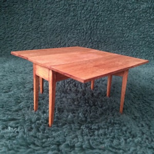 May include: A small wooden table with a drop leaf on each side. The table is made of light brown wood and has a simple design.