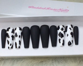 May include: Set of ten black and white cow print press-on nails. The nails are long and have a matte finish. The nails are arranged in a row on a white surface. The text "Studded Rose Nails" is visible in the background.