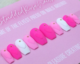 Puede incluir: Conjunto de uñas postizas de color rosa, blanco y rosa claro con un diseño de corazón y detalles dorados. Las uñas tienen forma de ataúd.