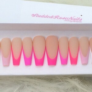 May include: Set of ten long, almond-shaped false nails with a white tip and a pink and hot pink ombre design. The nails are arranged in a row on a white surface. "Studded Rose Nails" is written on the surface.
