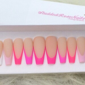 May include: Ten long, nude-colored, fake nails with a white tip and a pink or hot pink accent line. The nails are arranged in a row on a white surface.