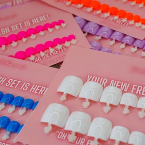 May include: Multiple sets of press-on nails arranged on pink display cards. Each card features the text "YOUR NEW FRESH SET IS HERE!" and "SITTING ON PRETTY". The nail sets come in various solid colors including neon pink, orange, purple, blue, and white.