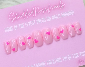 Puede incluir: Uñas postizas de color rosa con pequeños corazones rosas. Las uñas están dispuestas en una fila sobre un fondo rosa. El texto "Studded Rose Nails" y "Home of the Flyest Press On Nails Around!" es visible en el fondo.