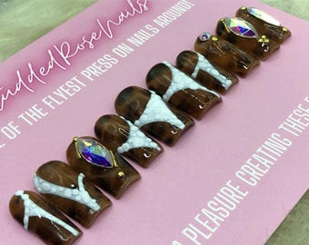 Puede incluir: Conjunto de diez uñas postizas marrones y blancas con un diseño abstracto blanco y pedrería. Las uñas se muestran sobre una superficie rosa con el texto "Studded Rose Nails" y "We of the flyest press on nails around!"
